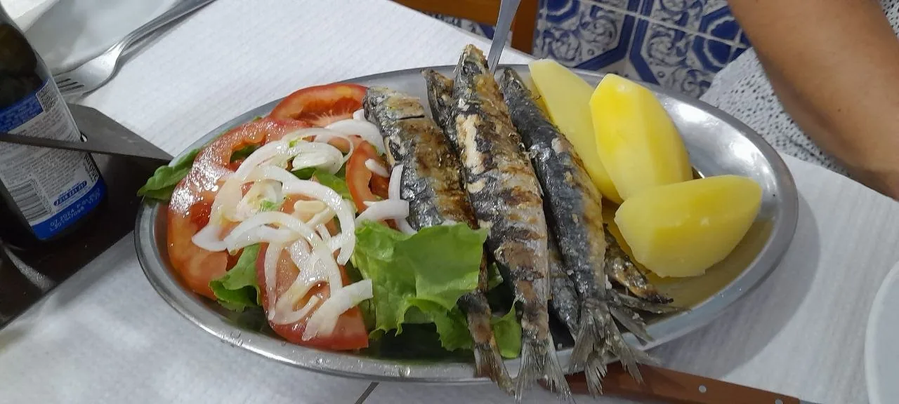 Een metalen schaal met gegrilde sardines, gekookte aardappelen en salade van tomaat en ui, tegen een muur met blauwe azulejos